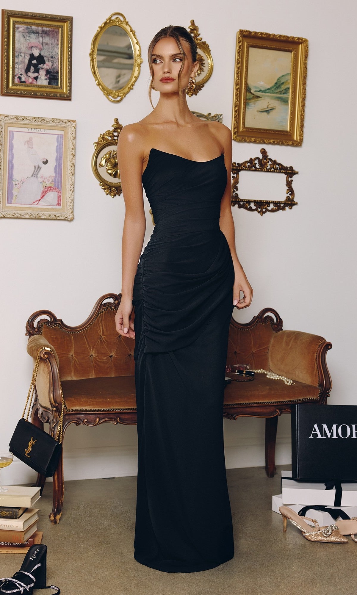 A woman in the black Amoris T726 long prom dress, featuring a strapless chiffon design and lace-up back, stands before a vintage-style sofa with framed art and decorative mirrors on the wall behind her.